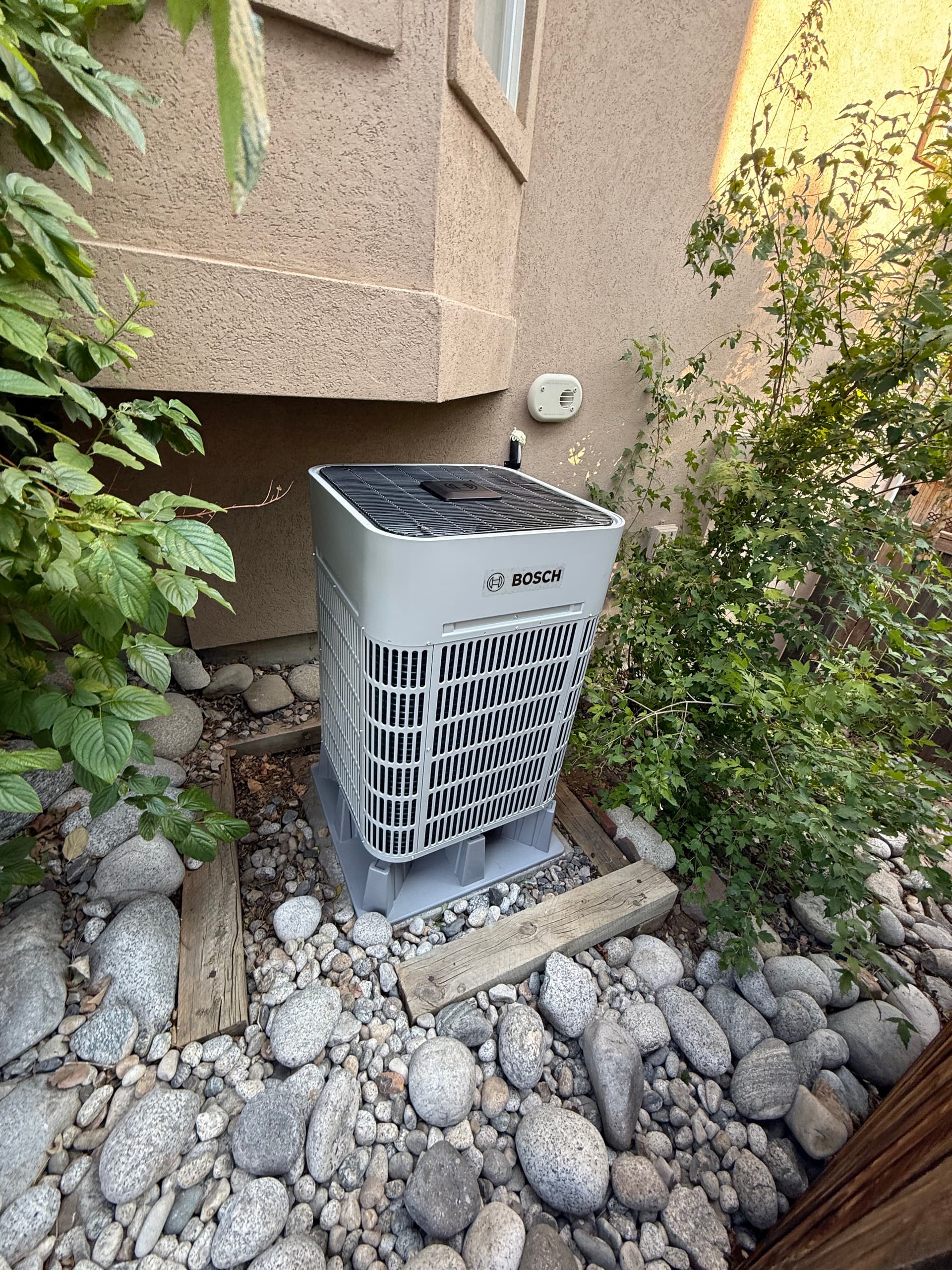Bosch cold climate heat pump outdoor unit on a rock bed beside a stucco home