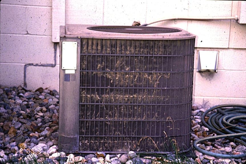 Dirty outdoor AC unit against gravel and brick wall with coiled refrigerant lines