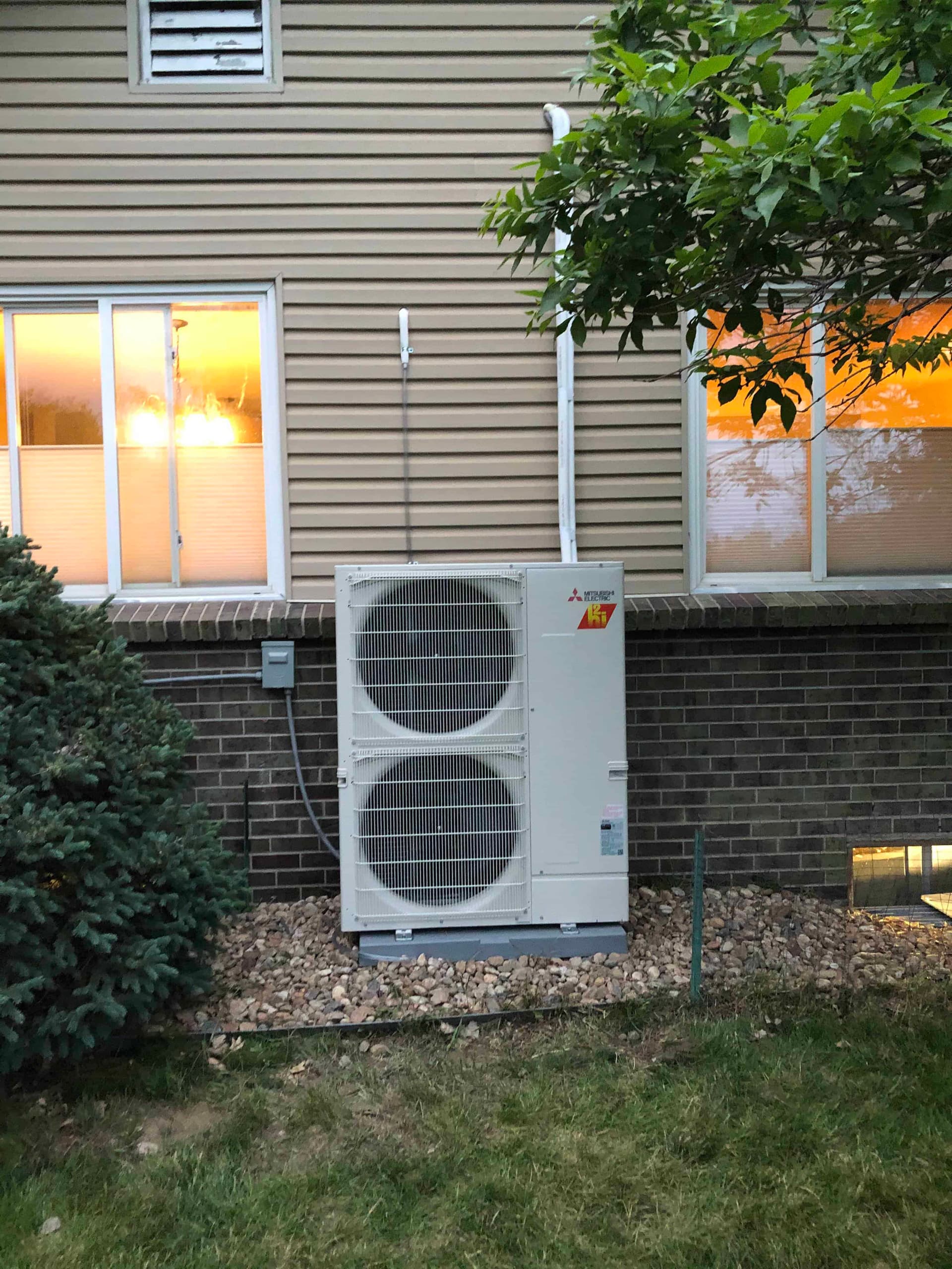 Mitsubishi heat pump outdoor unit installed beside brick wall with tree
