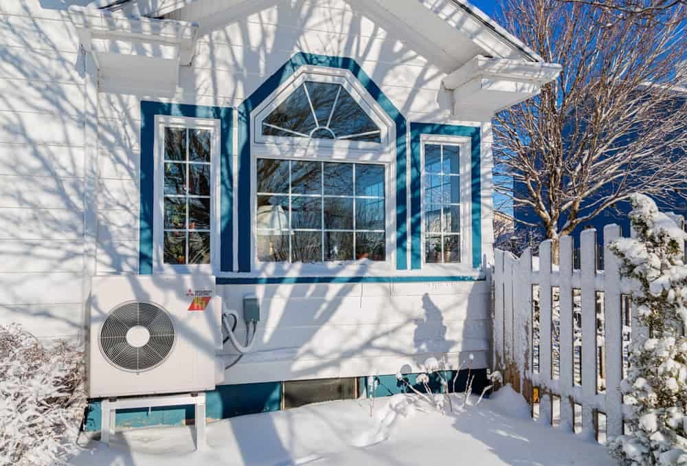 White house with blue trim and snow-covered yard, heat pump outdoor unit visible behind picket fence during Colorado winter