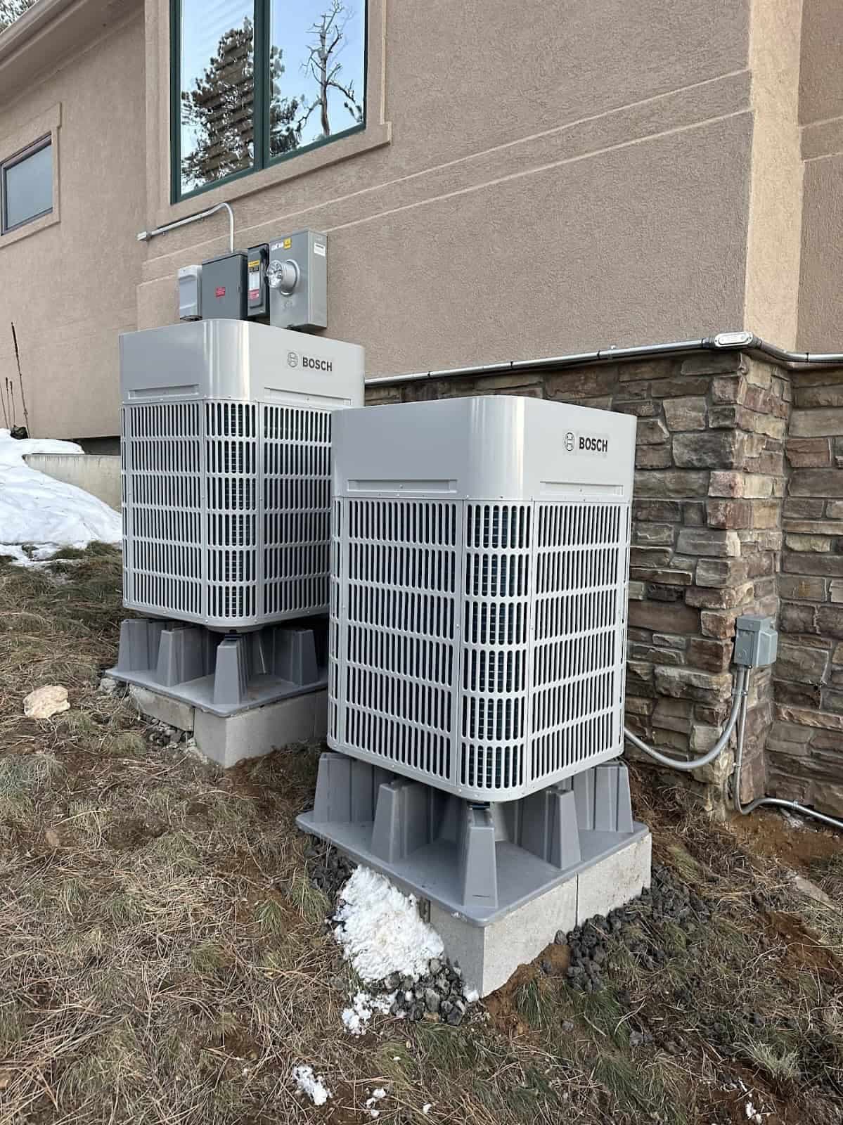 Two Bosch heat pump outdoor units on concrete pads beside a stucco and stone wall in winter, Colorado