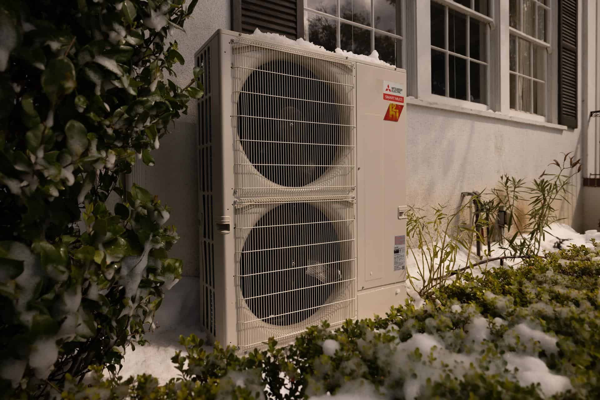 Mitsubishi Hyper-Heat two-fan outdoor unit running beside a house after a snowstorm