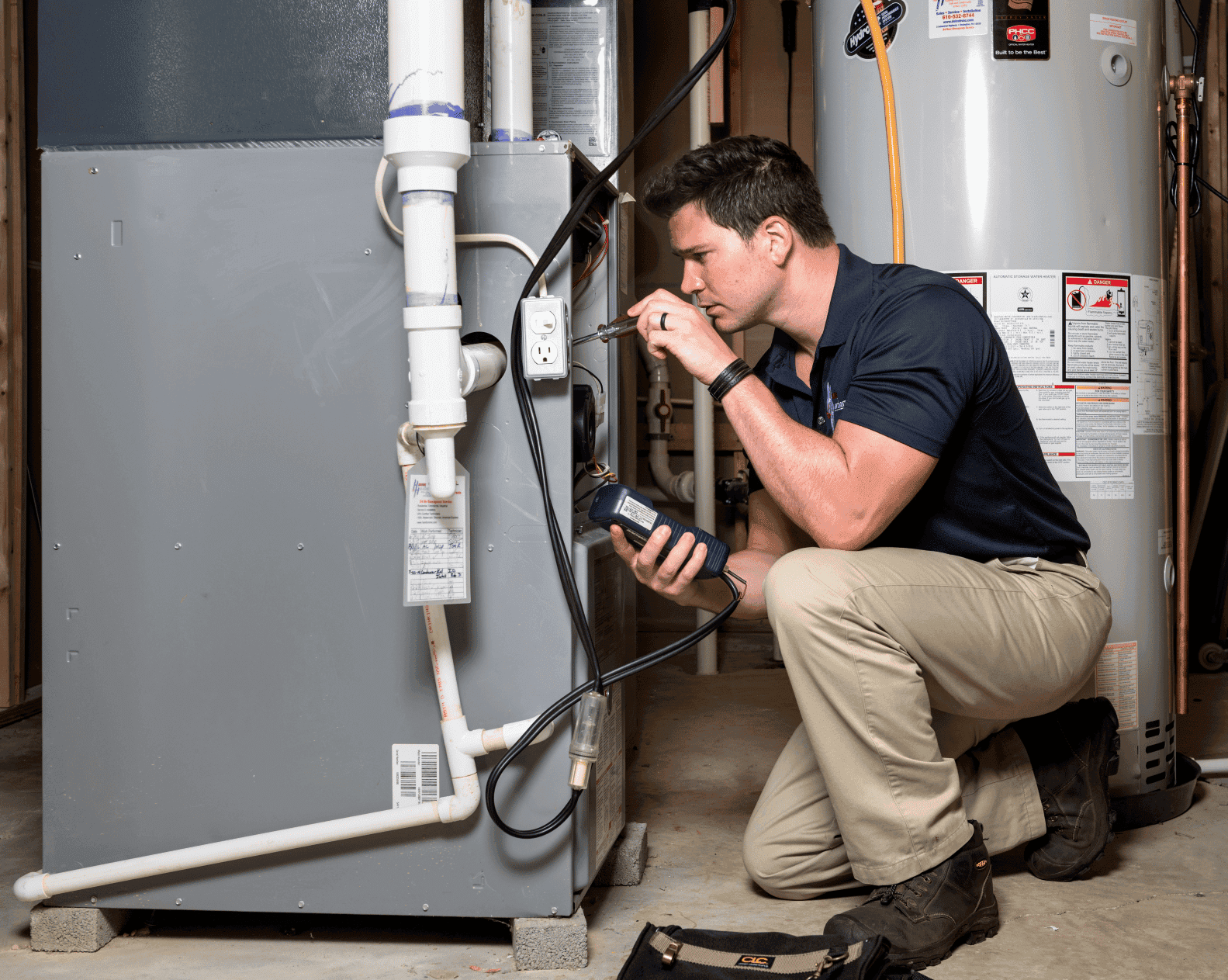 Technician using multimeter on gray furnace with white pipes, water heater with warning labels visible in utility room