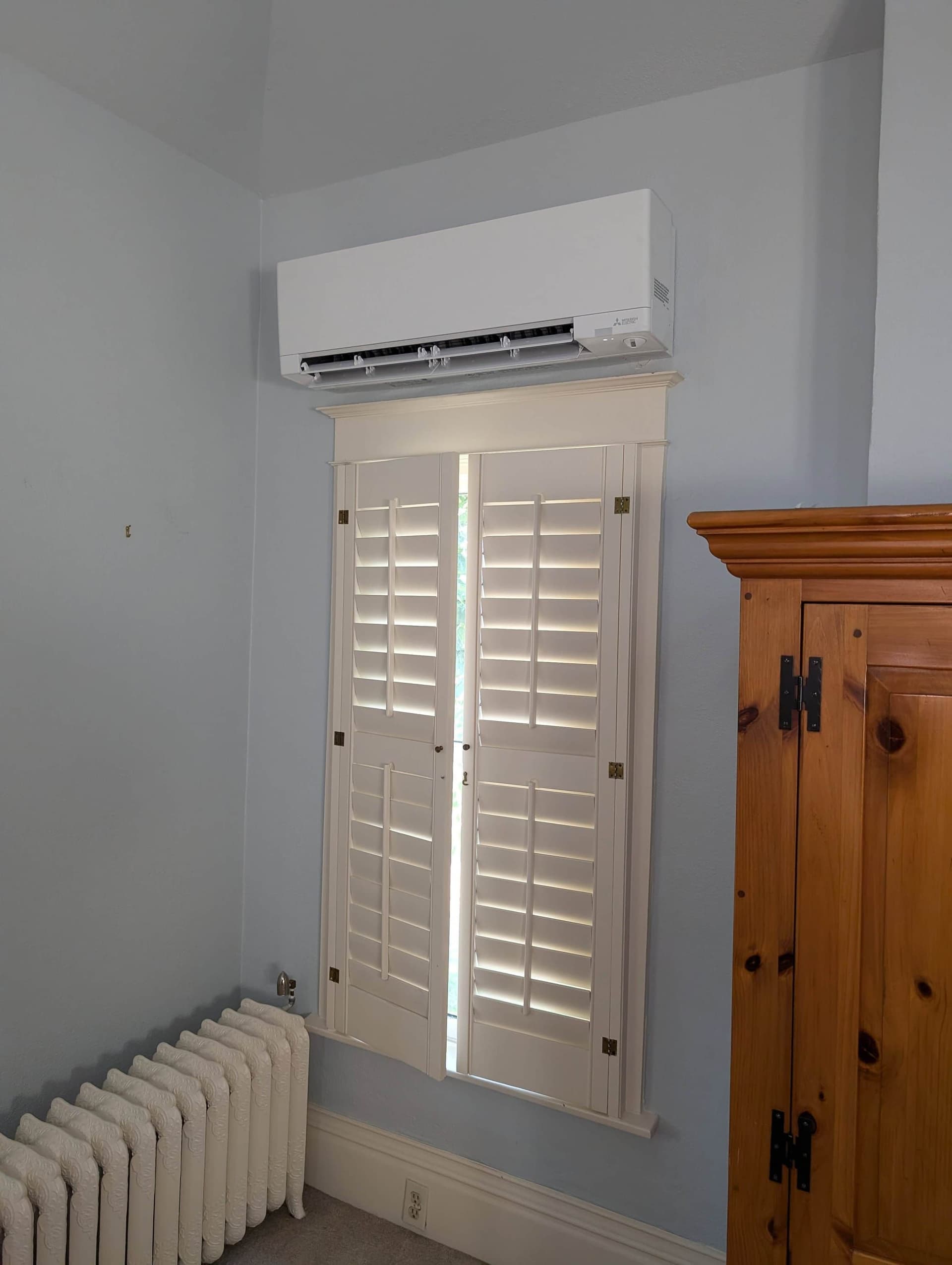 White mini-split indoor unit mounted above window shutters