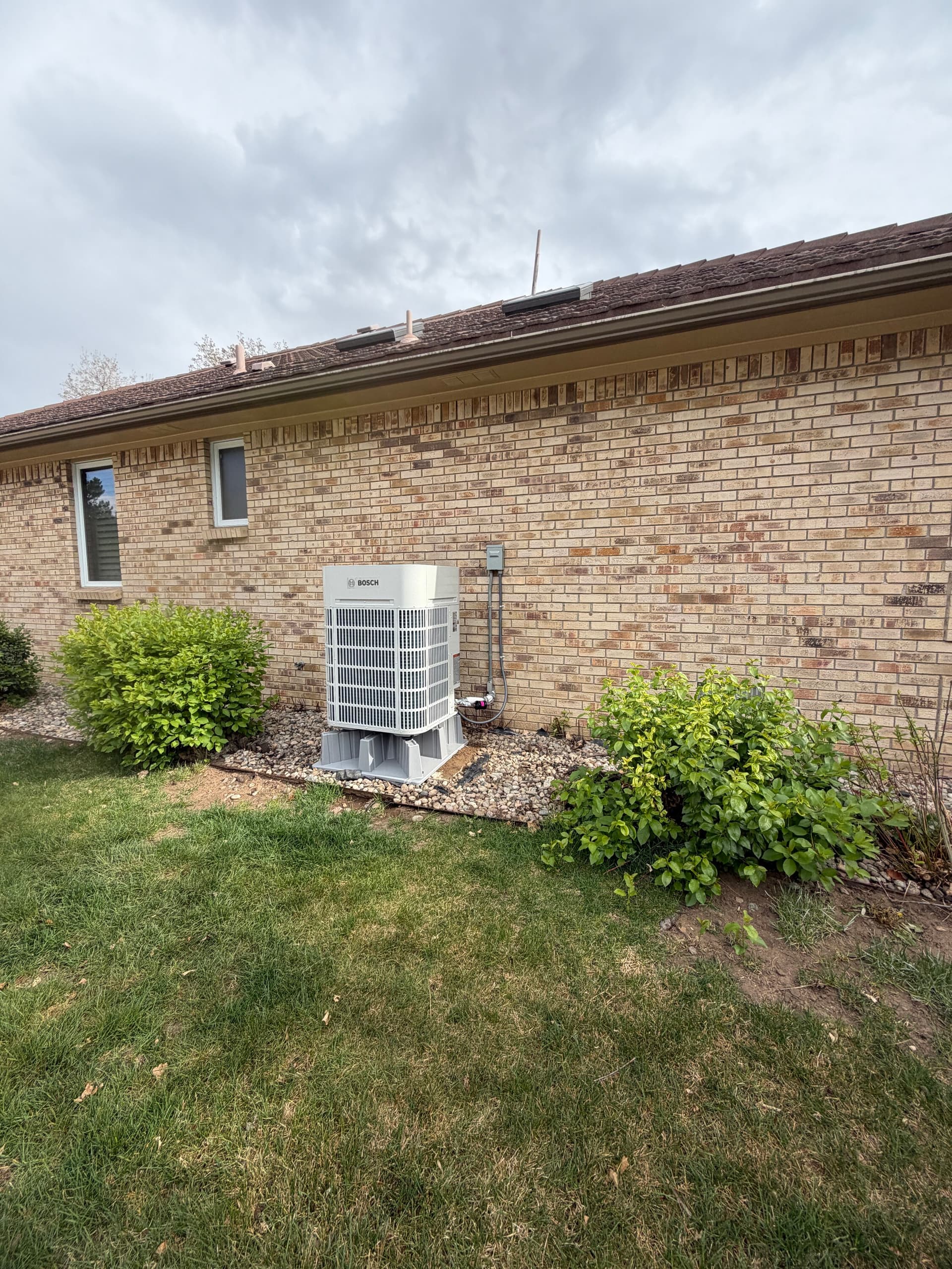 Bosch heat pump outdoor unit on a pad beside a tan brick ranch home in the Denver metro