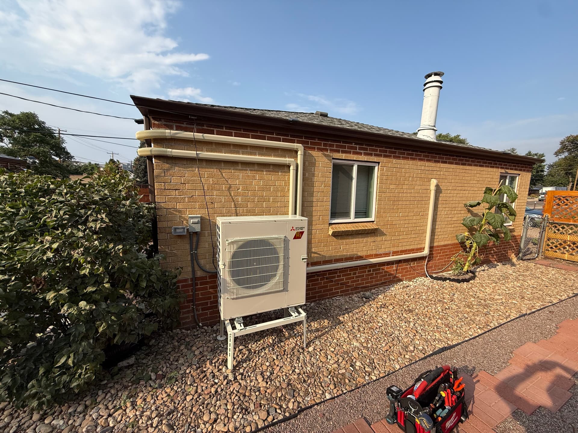 Mitsubishi heat pump outdoor unit on a stand beside a brick bungalow in a Denver neighborhood